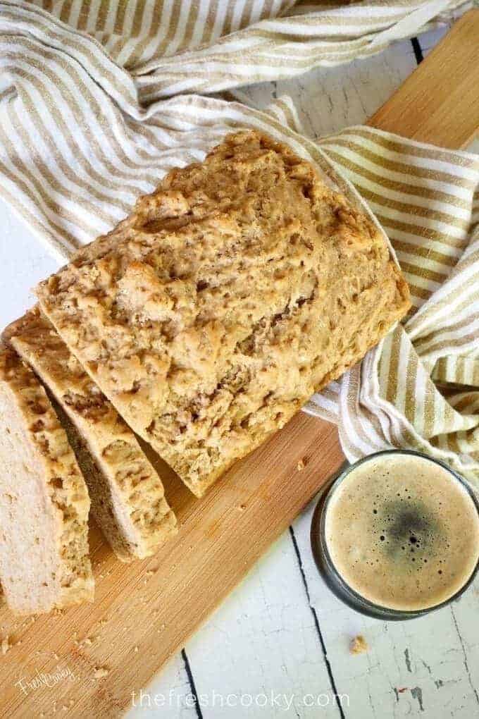 top down shot of a loaf of beer bread with two slices. A striped tea towel lies underneath and over a cutting board with a glass of dark beer in the foreground.