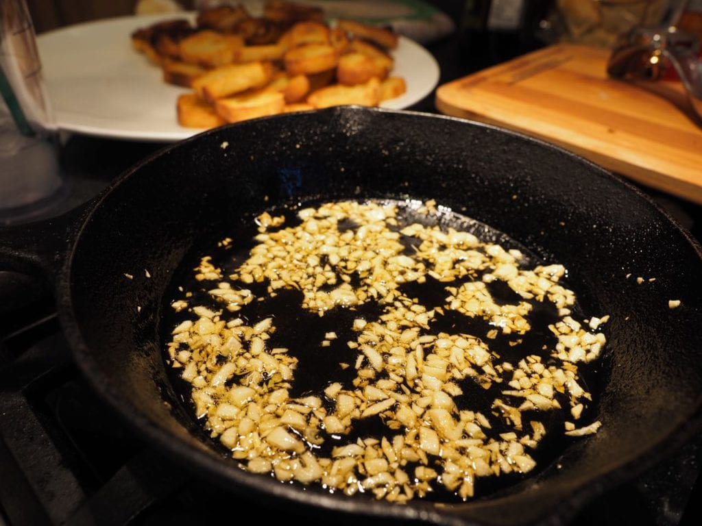 toasted garlic in olive oil in cast iron pan with toasted crostini in background.