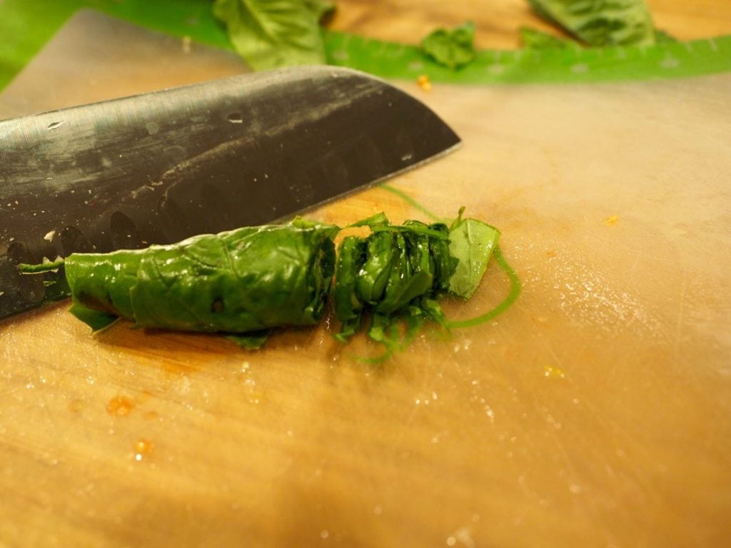 Rolled stack of basil leaves thinly sliced with knife in background. 