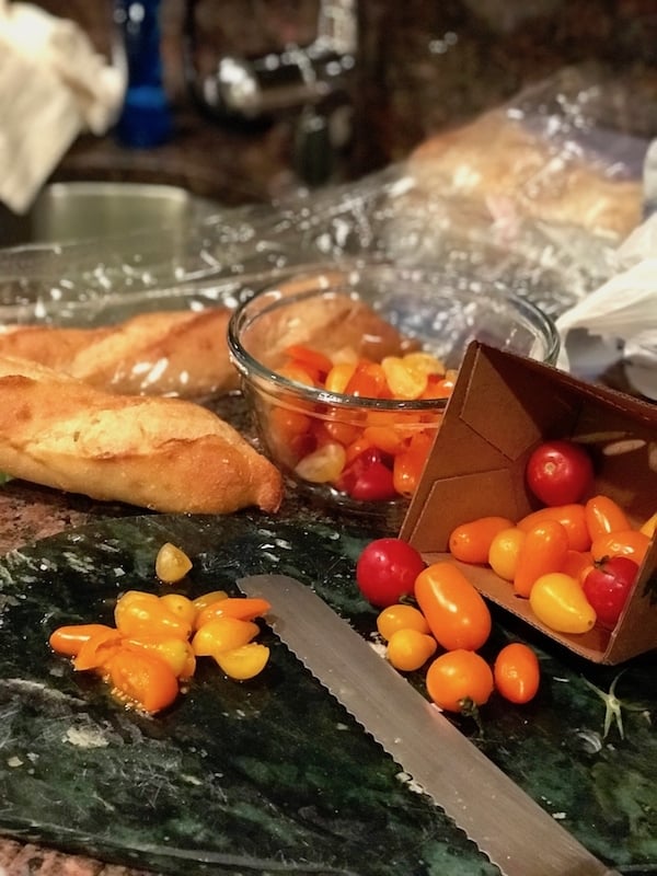 grape tomatoes in red, yellow and orange in a carton on a cutting board with a bowl of cut up tomatoes behind and a french baguette. 