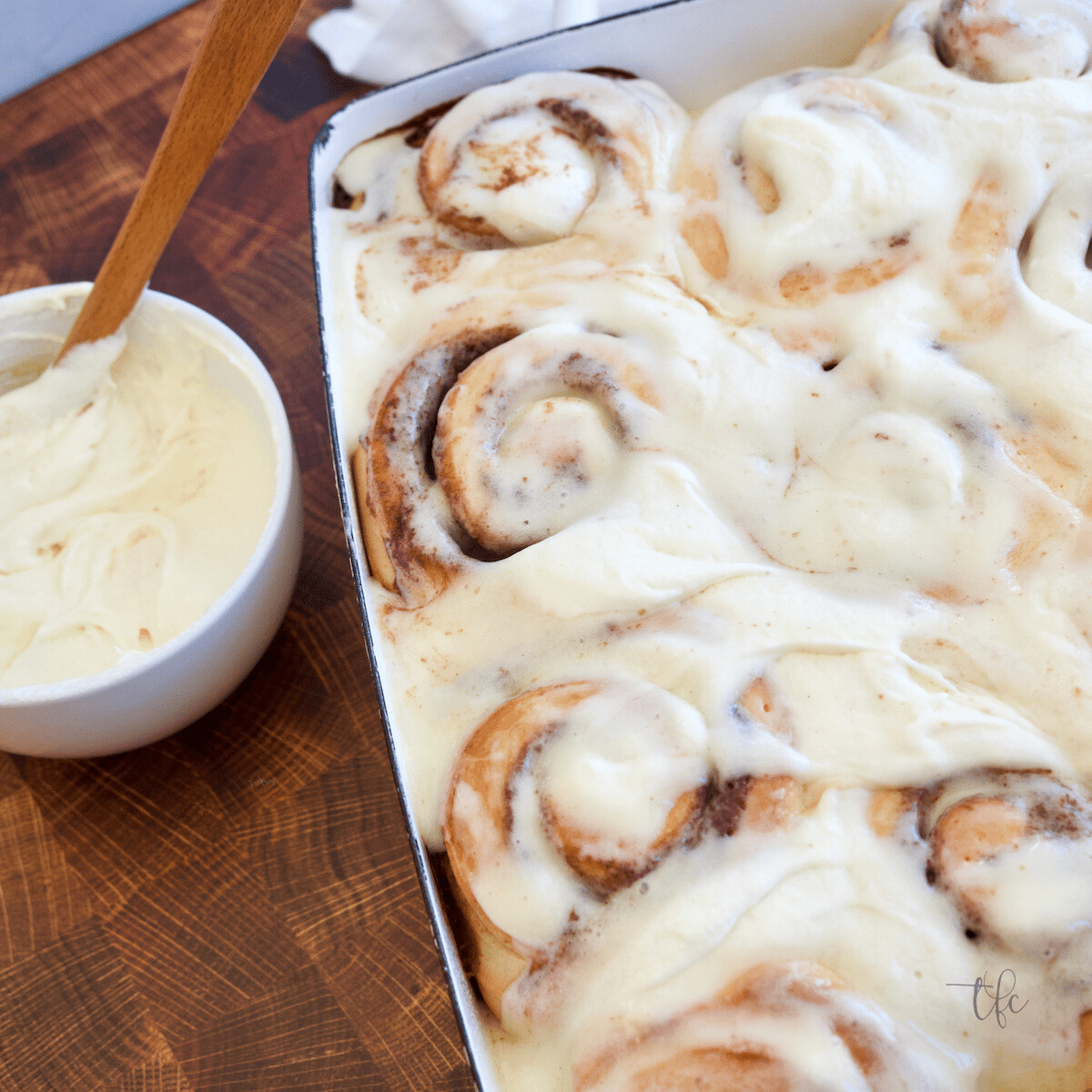 Ooey-gooey cinnamon rolls in a pan slathered with frosting with a bowl of frosting nearby.