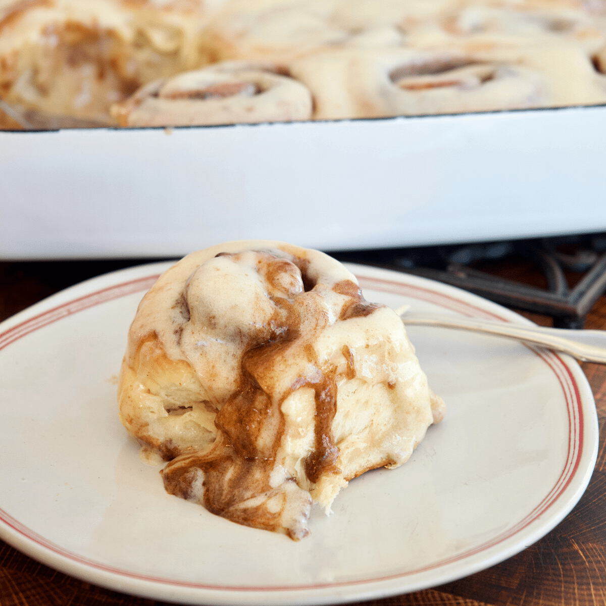 Single Gooey Cinnamon Roll on a plate with a tray of frosting cinnamon rolls behind.
