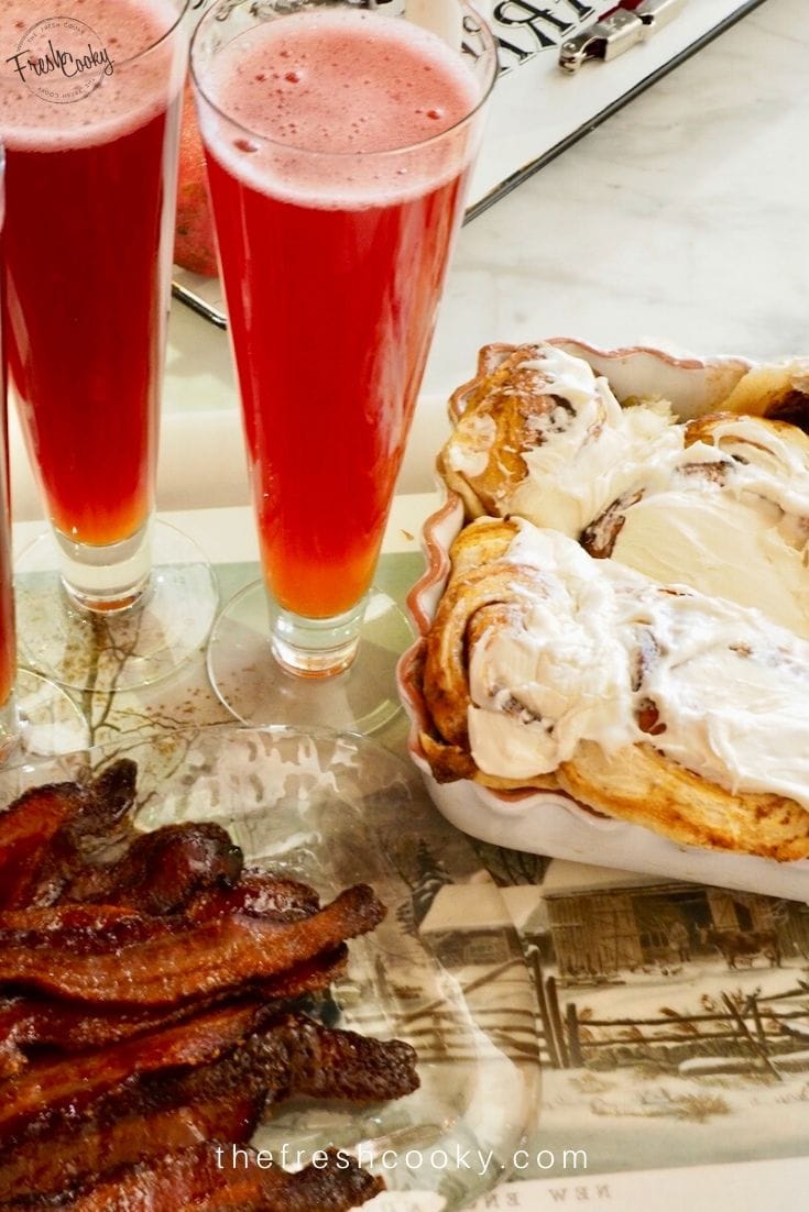 Cinnamon rolls with ooey cream cheese frosting on a tray with candied bacon and tall glasses of fizzy punch. 