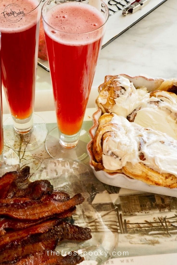 Cinnamon rolls with ooey cream cheese frosting on a tray with candied bacon and tall glasses of fizzy punch.