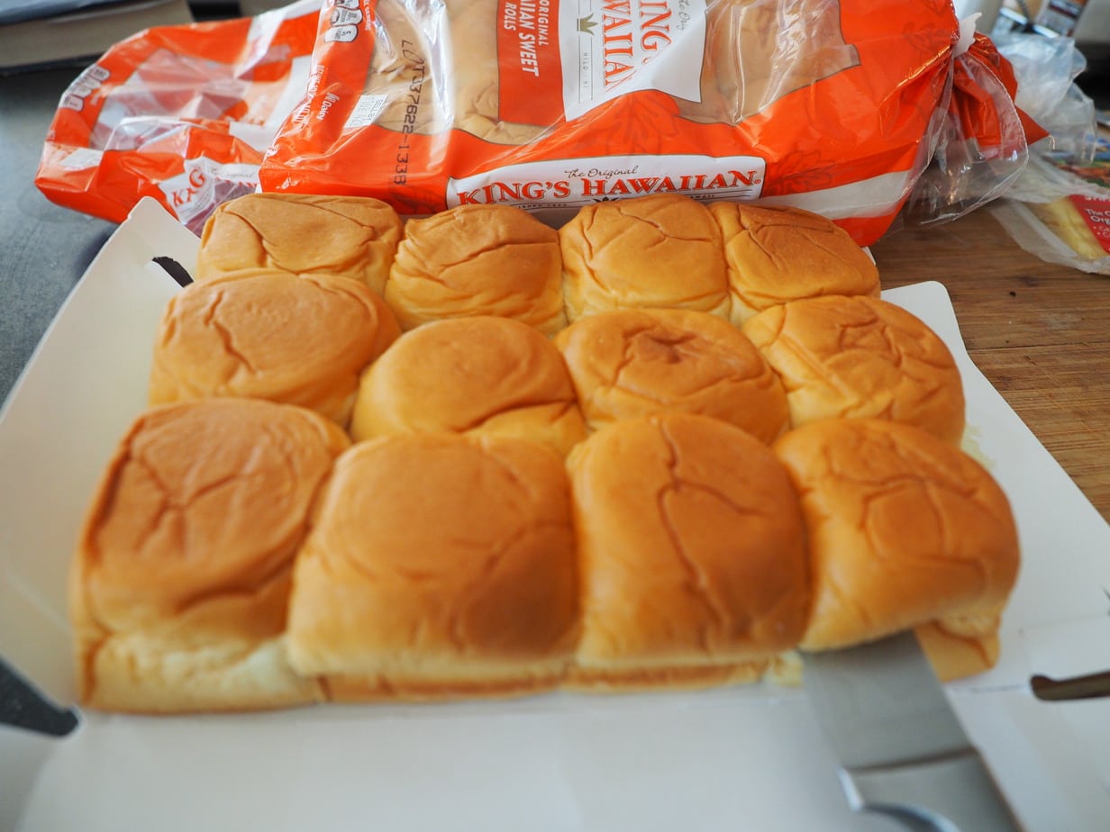 A tray of King's Hawaiian rolls on cutting board with knife slicing through. 