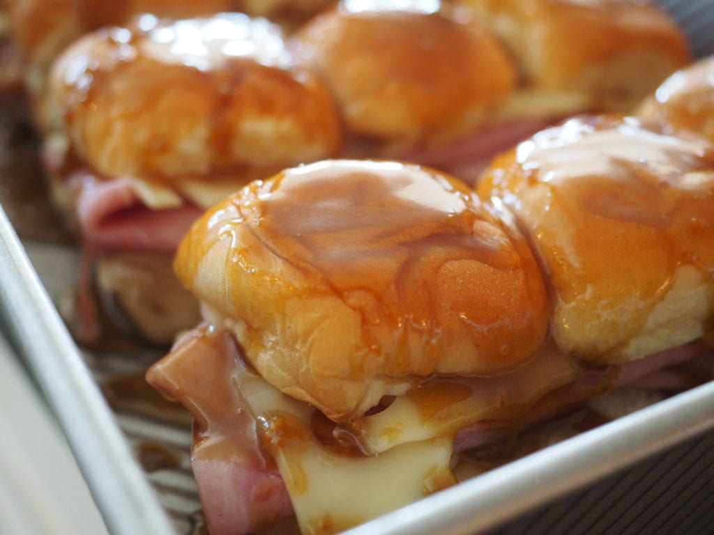 Close up shot of ham sandwiches in pan with sauce drizzled over tops and seeping down the sides. Ham & Swiss Sliders.