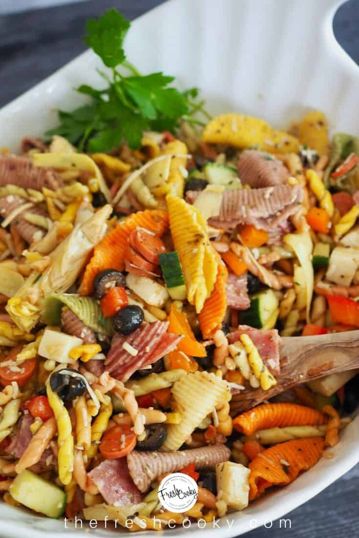 white bowl of tri-color twisty pasta salad with hunks of cheese, meatt, oliaves, artichoke hearts and basil.