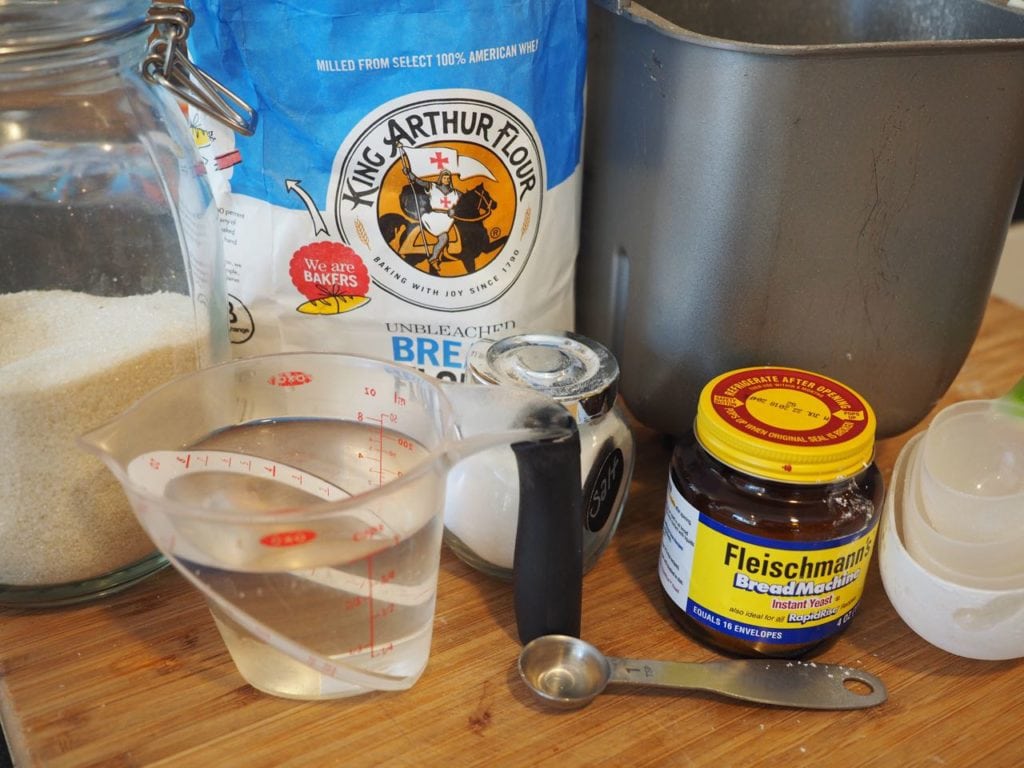 Ingredients for easy yeast rolls, left to right. Sugar, bread flour, bread machine, yeast, salt and water (plus egg, not shown)