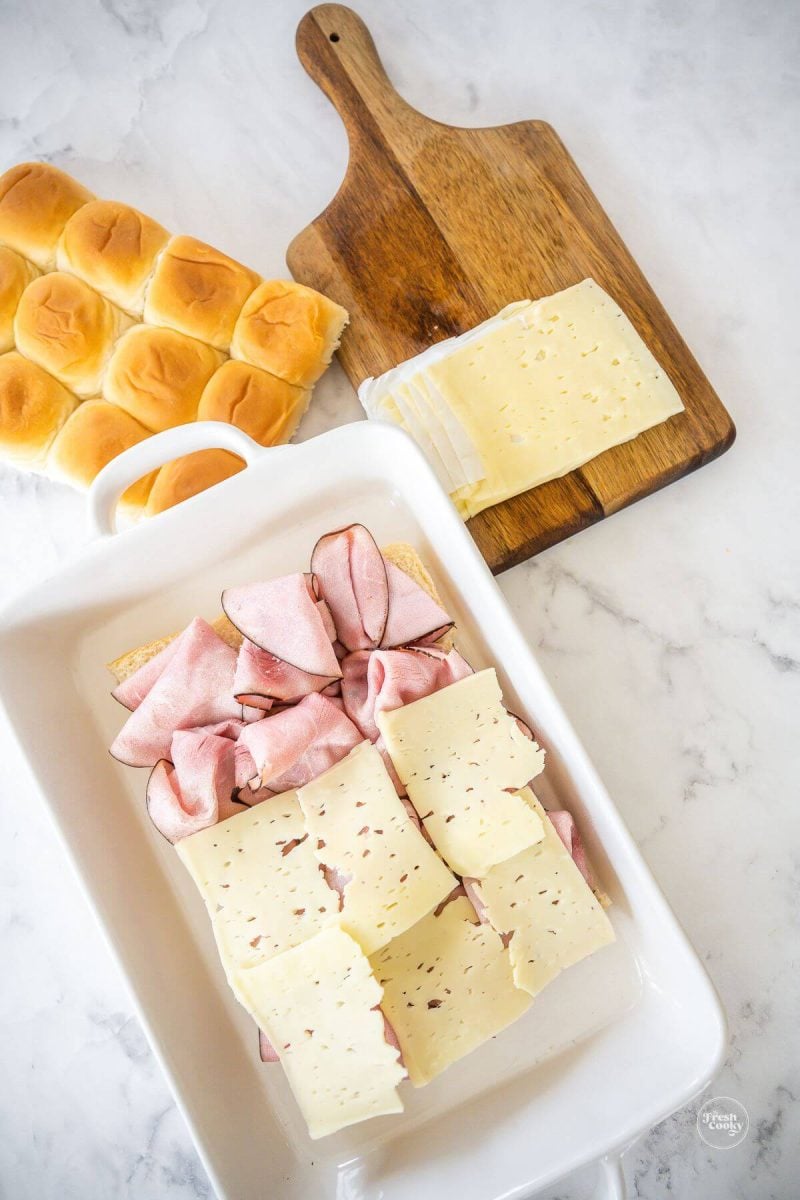Layering ham and swiss cheese onto sliced Hawaiian rolls.