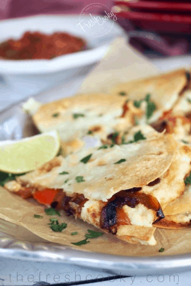 A silver tray with baked chicken stuffed quesadillas cut into triangles with a lime wedge and salsa in background. 