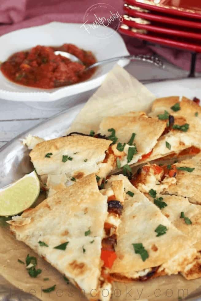 A silver tray with baked chicken stuffed quesadillas cut into triangles with a lime wedge and salsa in background.