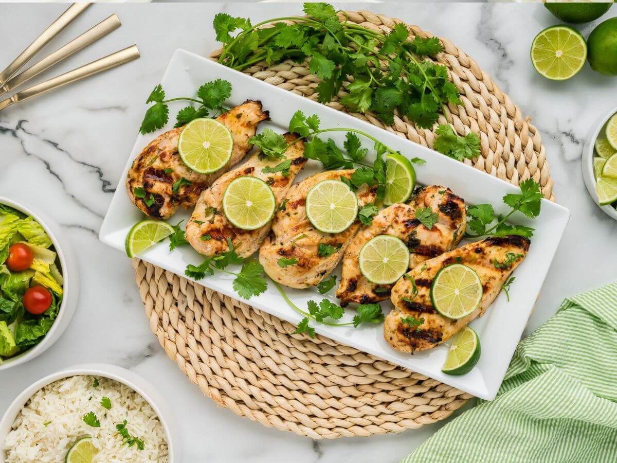 A round wicker placemat has a long serving plate of grilled, garnished chicken breasts next to salad and rice.