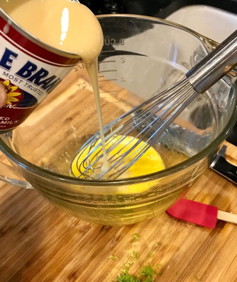 Pouring sweetened condensed milk into a glass bowl with a whisk over egg yolks. 