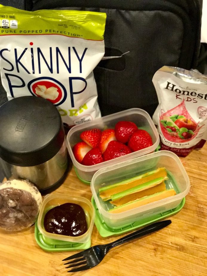 Black lunch box on cutting block with Thermos, rubbermaid containers filled with strawberries, celery with peanut butter, mini container filled with bbq sauce and a brownie sandwich cookie