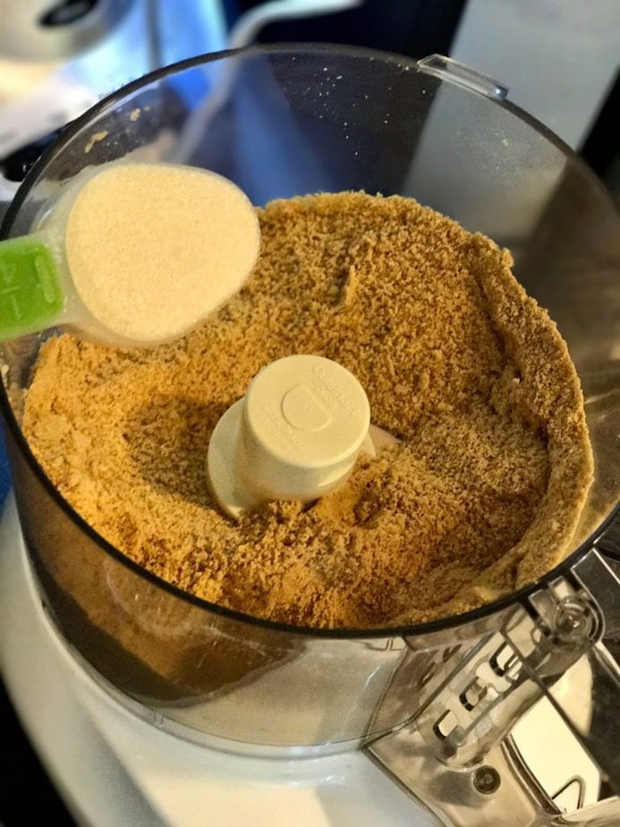 Graham cracker crumbs in food processor bowl pouring in some all natural sugar. 