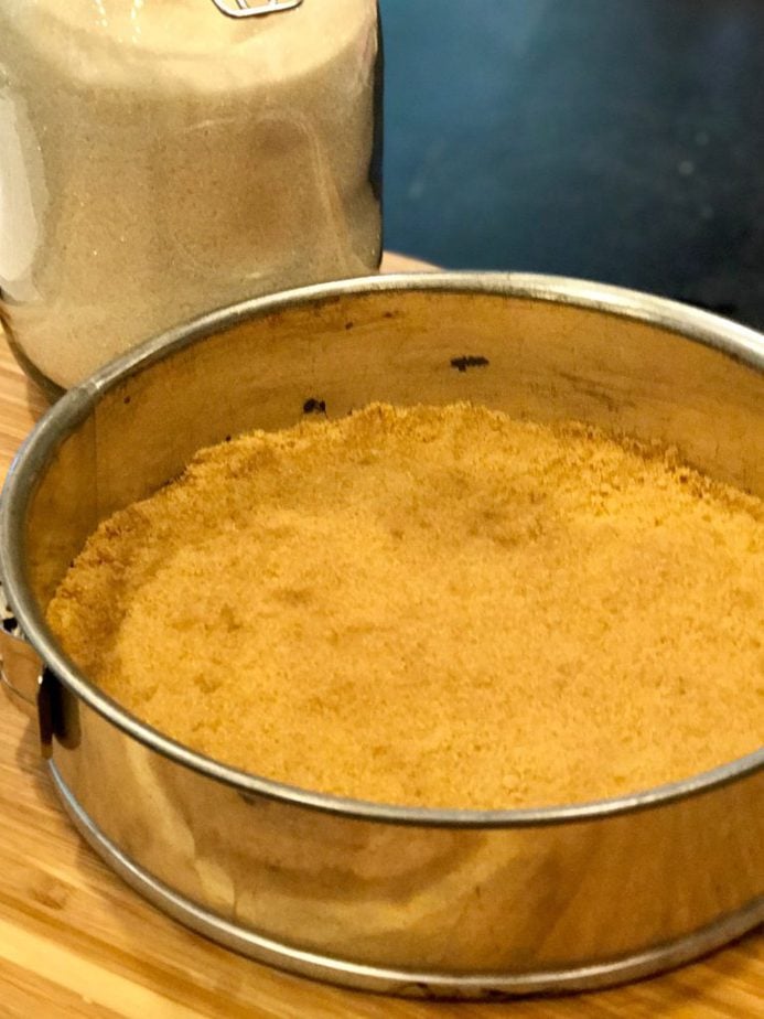 springform pan with graham cracker crumb crust pressed into bottom and slightly up sides of pan with a glass canister of all natural sugar in background. 