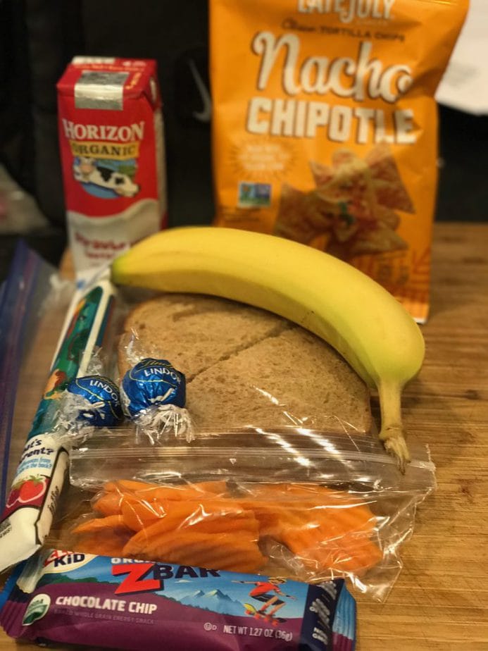 Traditional school lunch with lunch box in background, black Nike bag, with gogurt tube, ham sandwich, banana, carrot chips, Z Bar and two chocolate lindt balls, plus a strawberry milk and Nacho Chipotle Chips