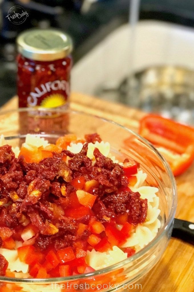 Jar of sun-dried tomatoes in background and another poured on top of the pasta salad. 