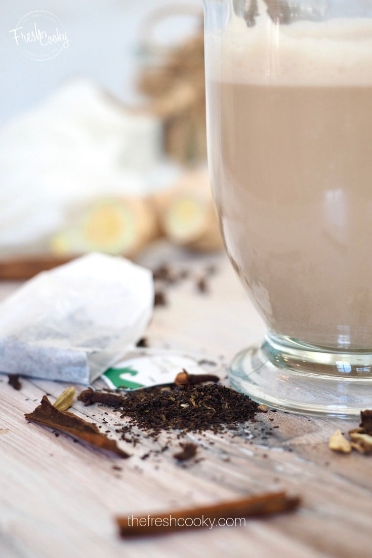 Ingredients for Chai Tea Latte and mug of latte with tea bag, cinnamon stick, cloves and cardamom strewn on the table www.thefreshcooky.com