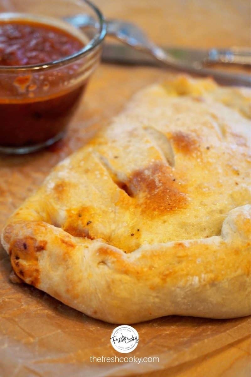 Baked Easy Calzone on parchment paper with fork and knife in background and dipping sauce to the side