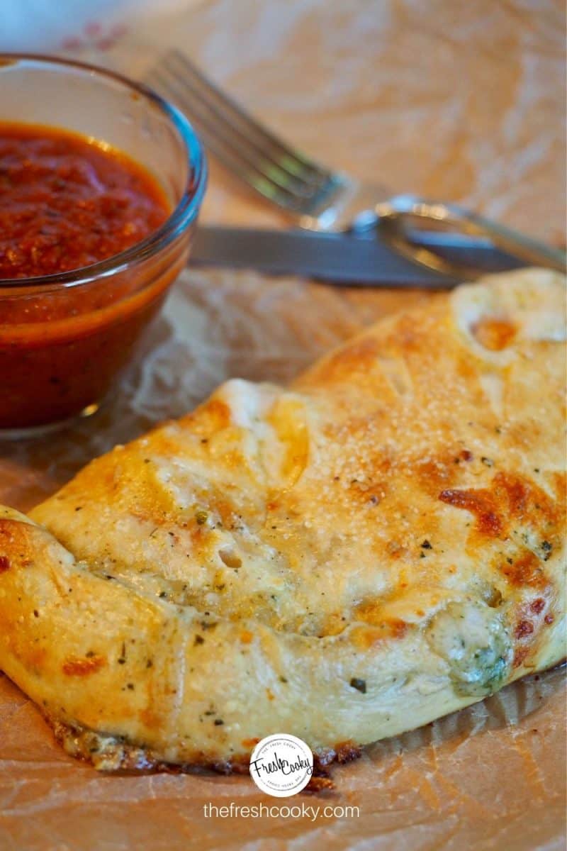 Baked Easy Calzone on plate with parchment paper, dipping tomato sauce in background with fork and knife.