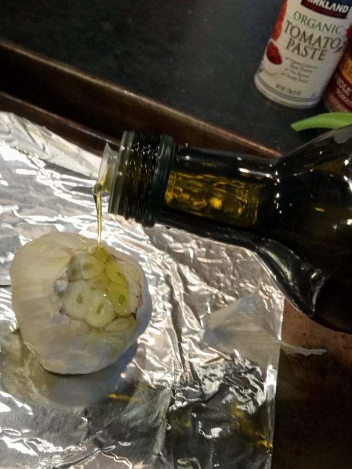 Pouring olive oil over the cut top of a bulb of garlic. 