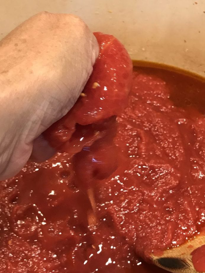 Hand squeezing whole tomatoes into the spaghetti sauce. 