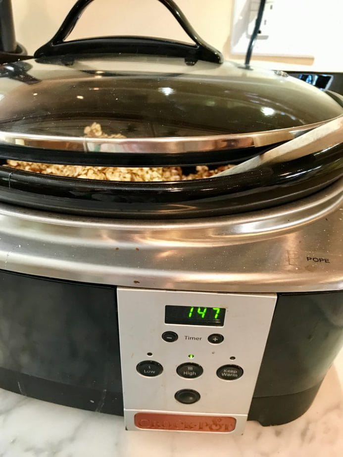 CROCKPOT with granola inside, a wooden spoon sticking between the lid and the crock to vent the steam. 