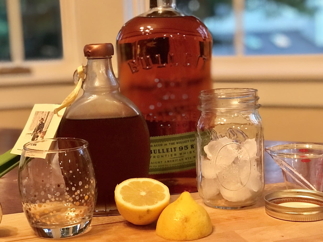 Ingredients for maple whiskey sour. Left to rightbourbon maple syrup, Bulleit Rye Whiskey, a mason jar with ice in it, a mini measuring cup, a cocktail glass with gold spots and a sliced lemon all sitting on a cutting board. 