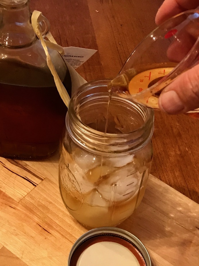pouring maple syrup into mason jar over ice on cutting board using a mini liquid measuring cup, with maple syrup jar sitting off to the left back. 