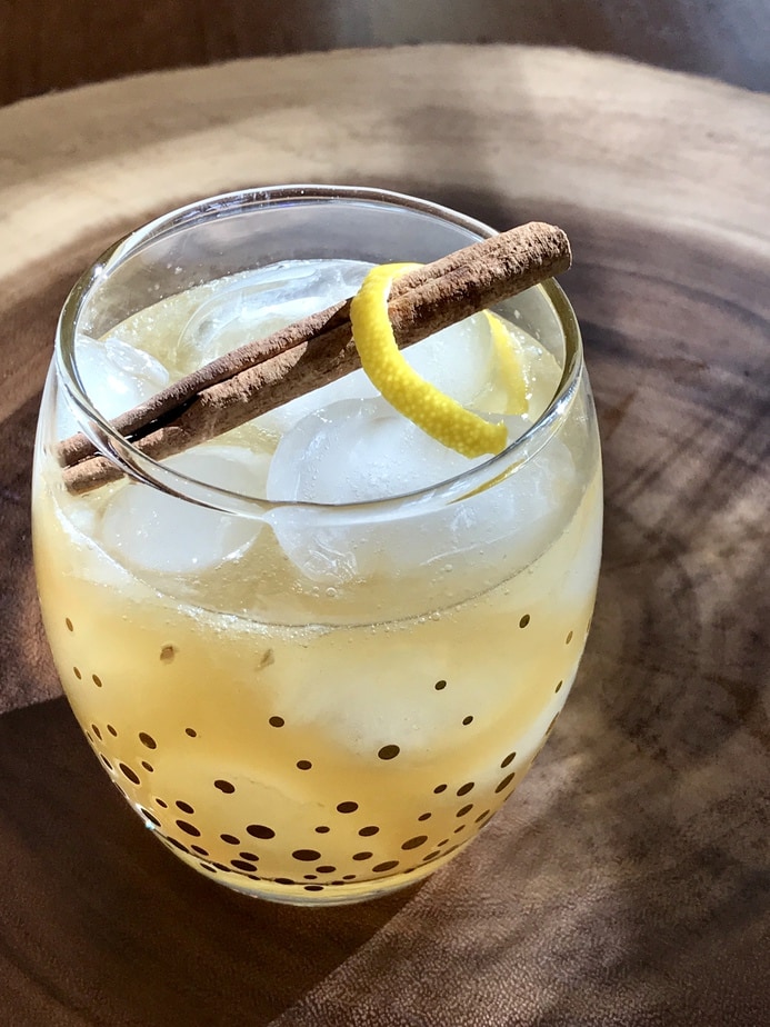Maple Whiskey Sour in a glass, sitting on wooden round lazy suzan, the light streaming in just right to highlight the cinnamon stick and lemon curl. 