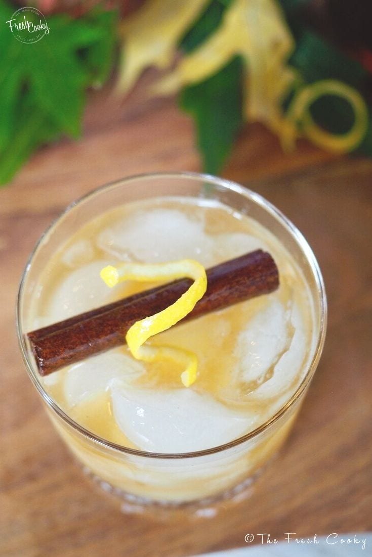 Closeup shot of whiskey sour with cinnamon stick and lemon rind swizzle on a cutting board with leaves in background. 