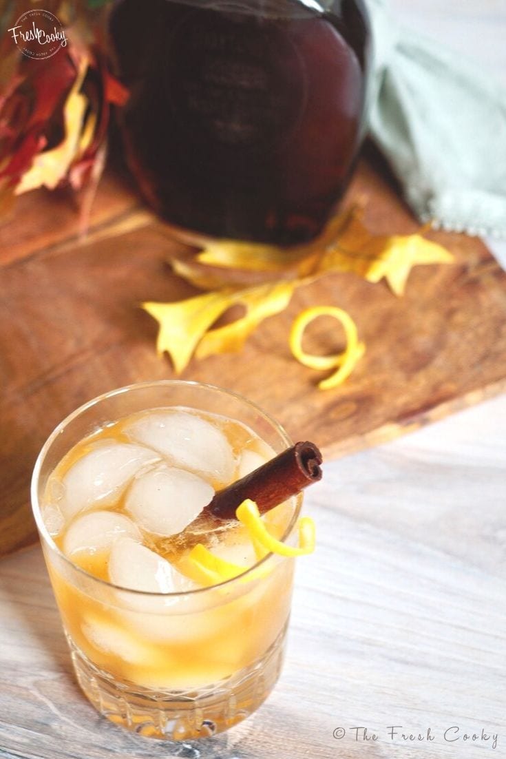 Cocktail glass with ice cubes and filled with Maple Whiskey Sour with Cinnamon stick and lemon swizzle of rind stick out side of glass, on cutting board with a hickory cutting board in background and golden maple leaf. 
