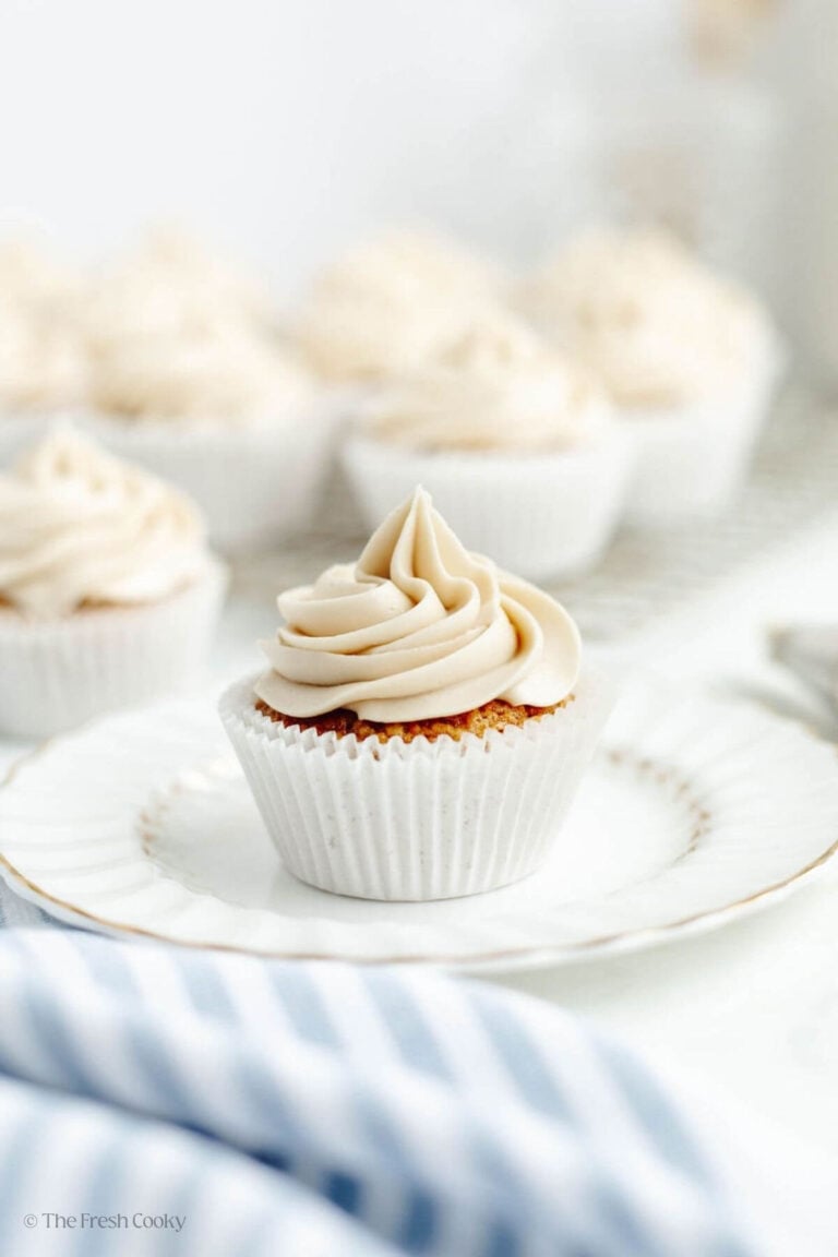 A single double lined carrot cake cupcake frosted with a thick swirl of frosting on a pretty plate, with more cupcakes behind. 