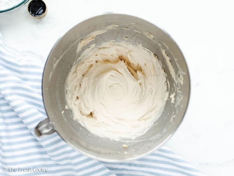Cream cheese ingredients in mixing bowl of stand mixer, with streaks of brown from adding vanilla and brown sugar.
