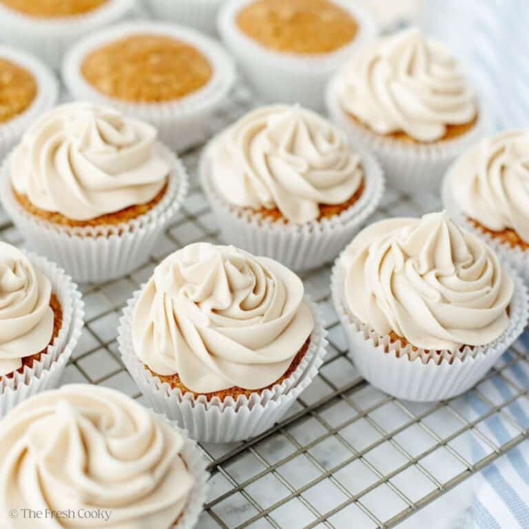 Piped and frosted carrot cake cupcakes on a wire rack, with some unfrosted cupcakes in background.