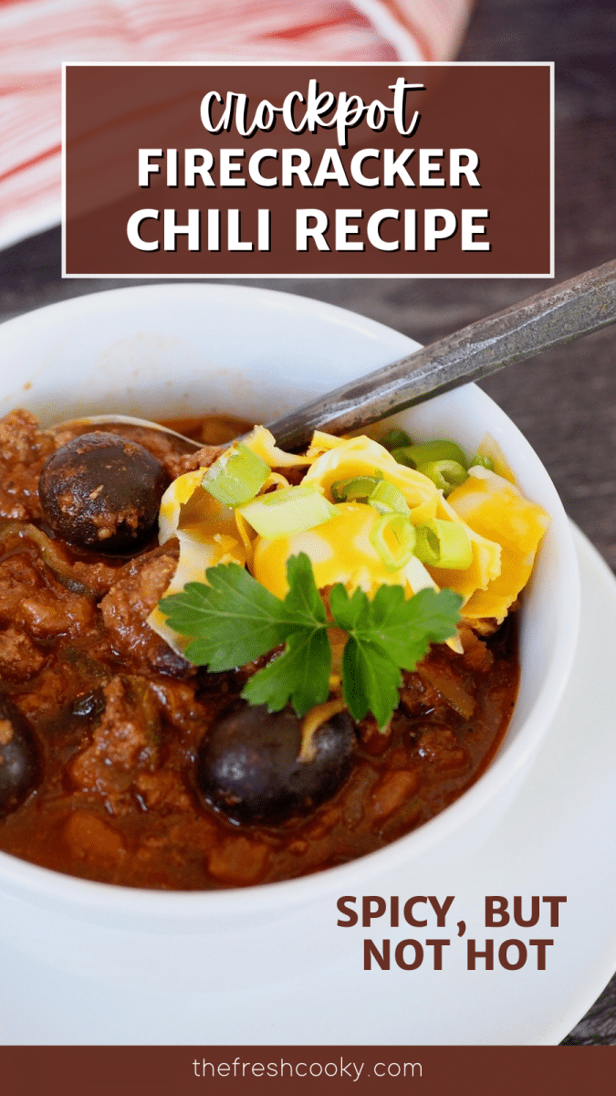 Pin for crockpot firecracker chili recipe with white bowl filled with rich olive studded firecracker chili with shredded cheddar cheese and a spoon.
