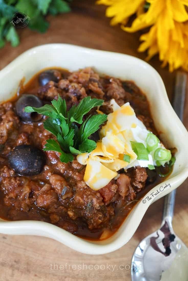 bowl of firecracker chili with shredded cheese, parsley and black olives