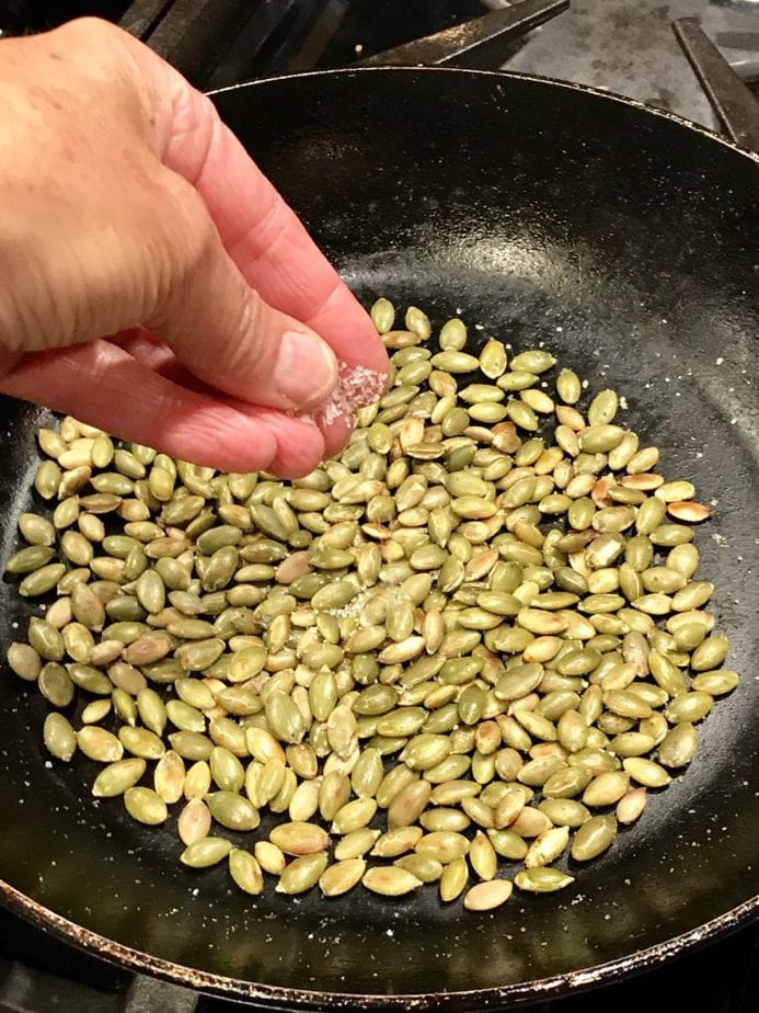 Hand adding a pinch of salt to toasted pumpkin seeds in fry pan. 