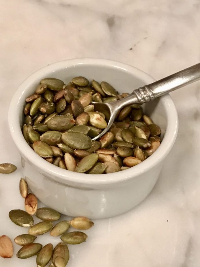 Close up of toasted pumpkin seeds in a small white ramekin with mini silver spoon sitting on marble. 