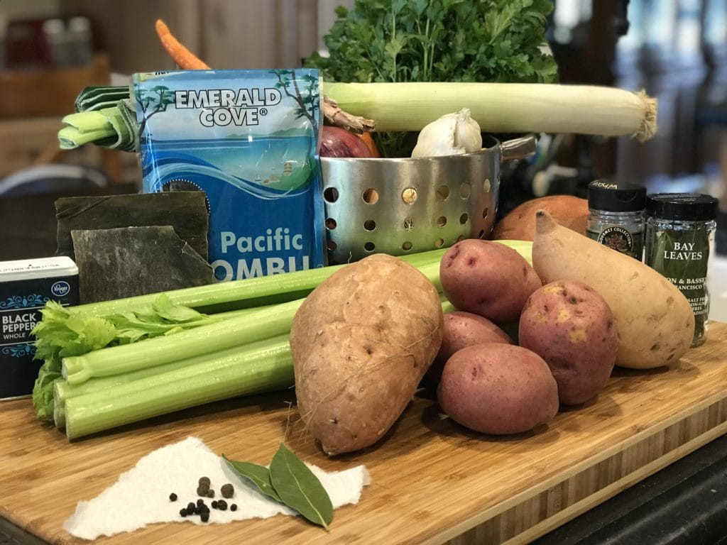 Ingredients for Magic Mineral broth, left to right black pepper, kombu seaweed, colander with leek, onions, parsley, bay leaves, sweet potatoes, baked potatoes. 