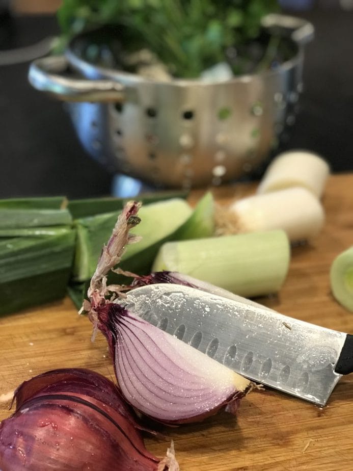 Cutting board with knife slicing, a red onion, leeks chopped rough in back. 
