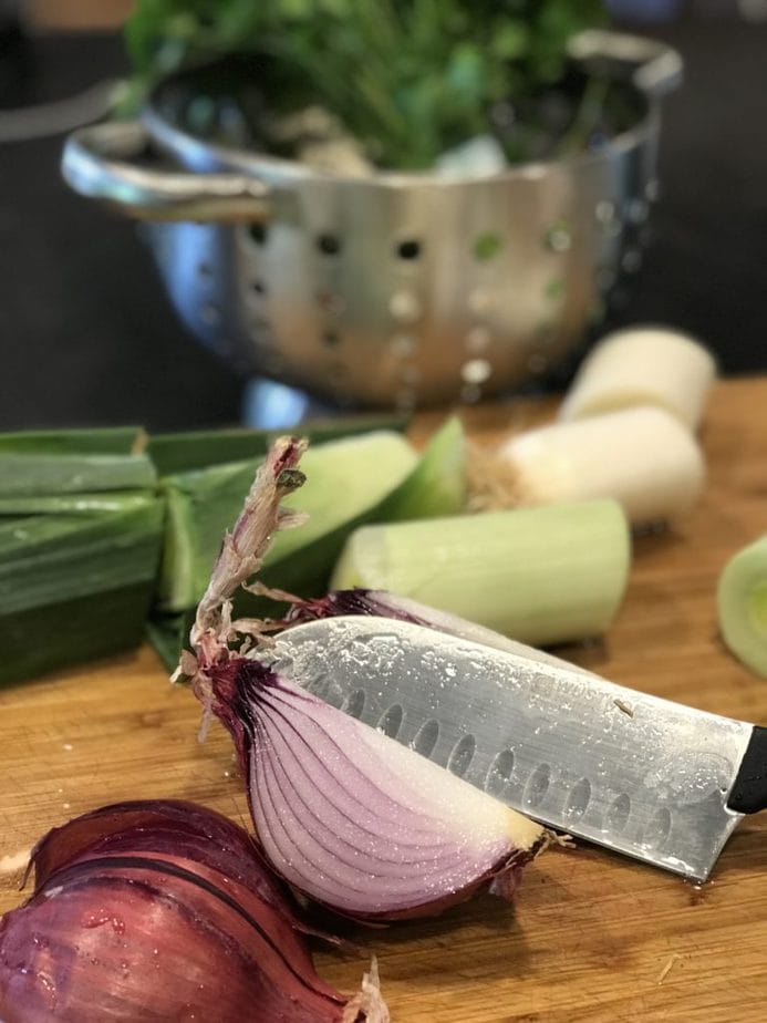 Cutting board with knife slicing, a red onion, leeks chopped rough in back.