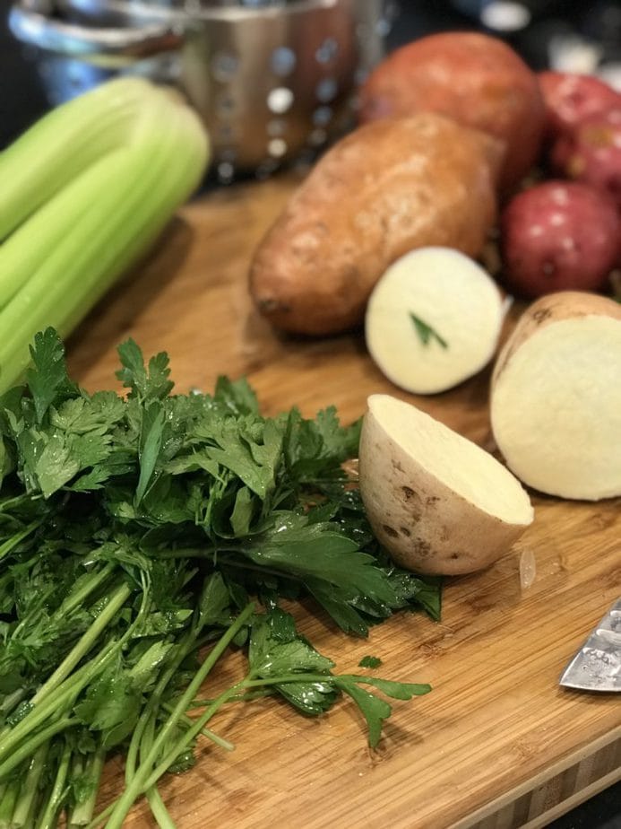 cutting board with parsley, celery, chopped potatoes and washed sweet potatoes and red potato in the background. 