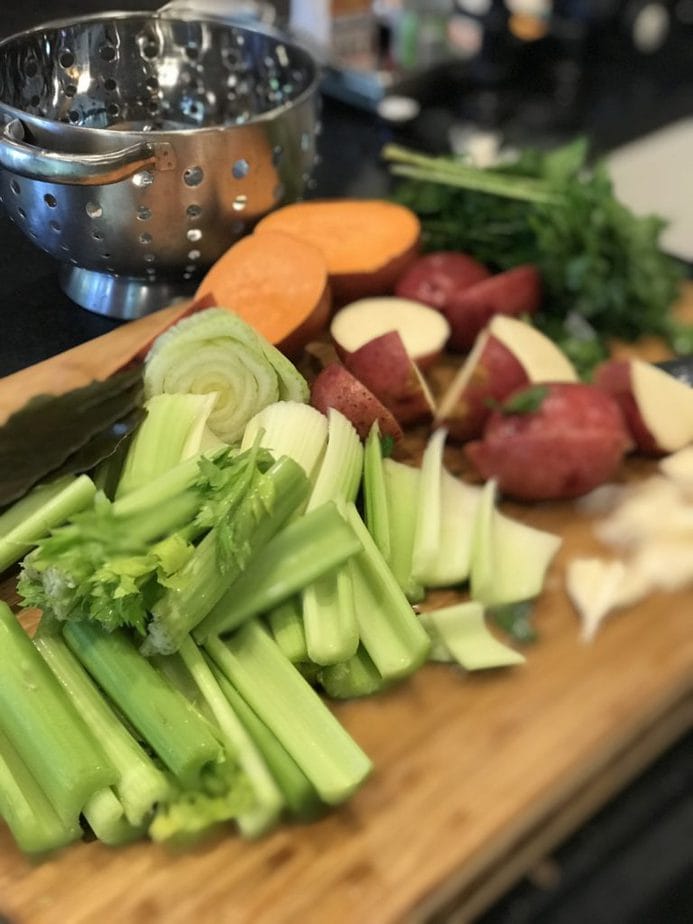 Cutting board with large slices of celery, potatoes, sweet potatoes and garlic with a colander in the background. 