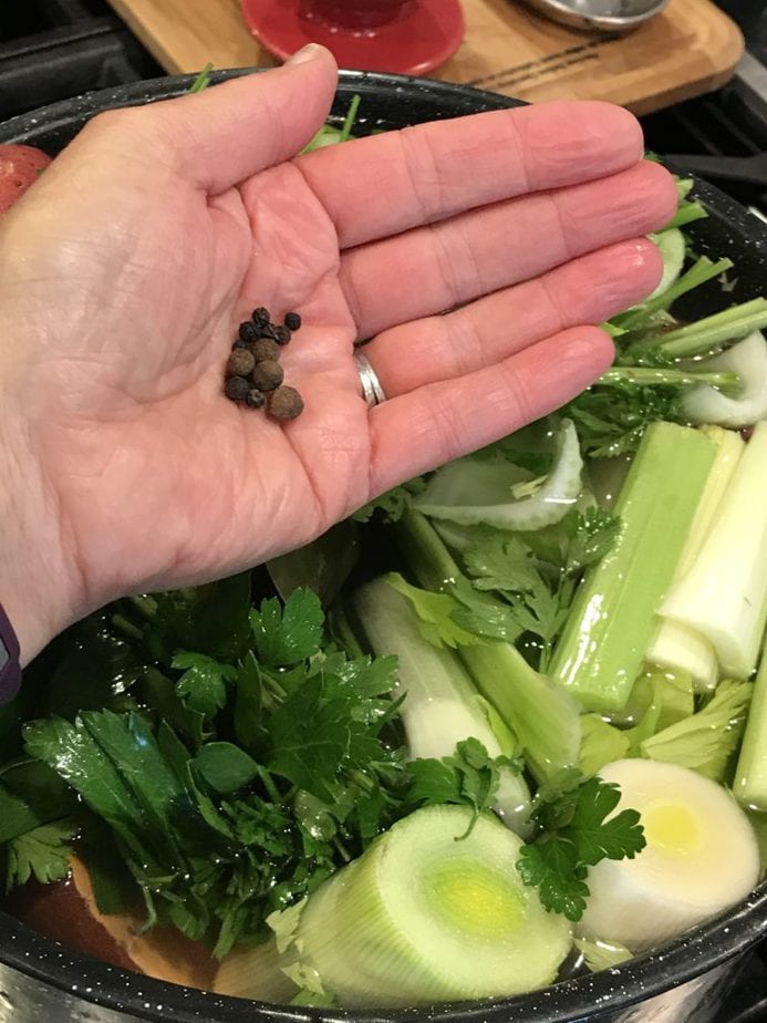 all spice and black peppers in palm of hand over pot of water with chopped veggies