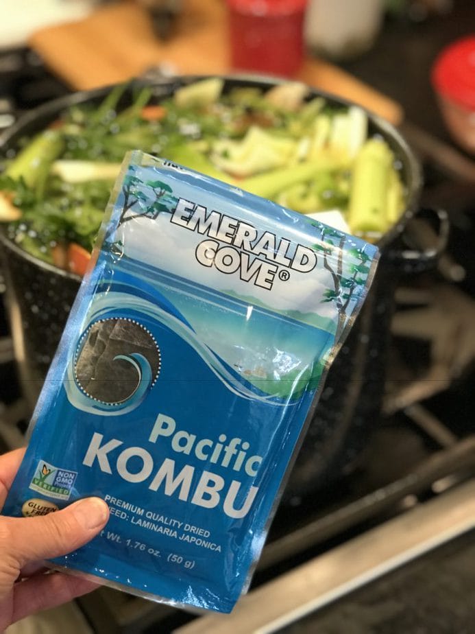 Hand holding a package of Emerald Cove Pacific Kombu seaweed in front of large pot filled with water and veggies.