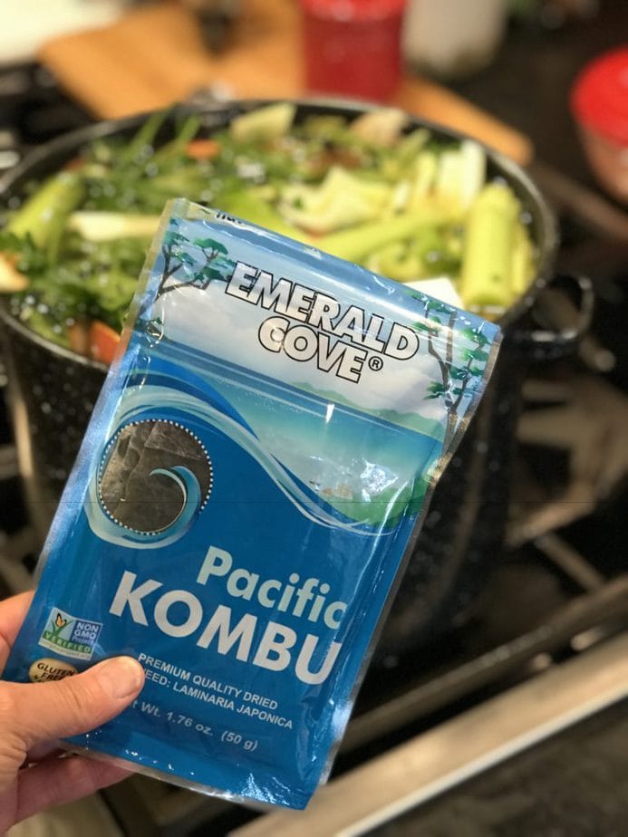 Hand holding a package of Emerald Cove Pacific Kombu seaweed in front of large pot filled with water and veggies. 