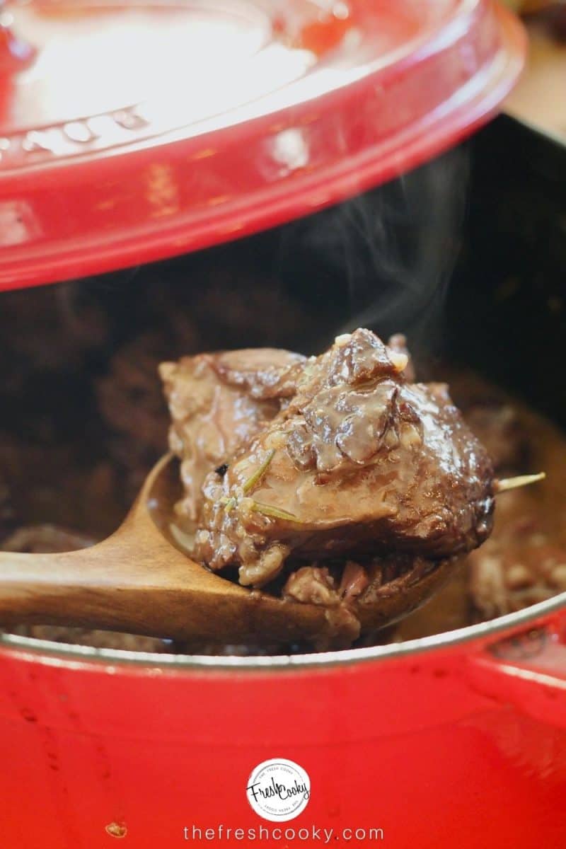 Steaming hunk of pot roast coming out of a red dutch oven on a wooden spoon
