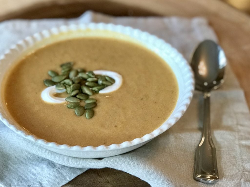 A bowl of creamy orange pumpkin soup garnished with pumpkin seeds and sour cream on a wooden board with a linen napkin and spoon resting beside the bowl. 